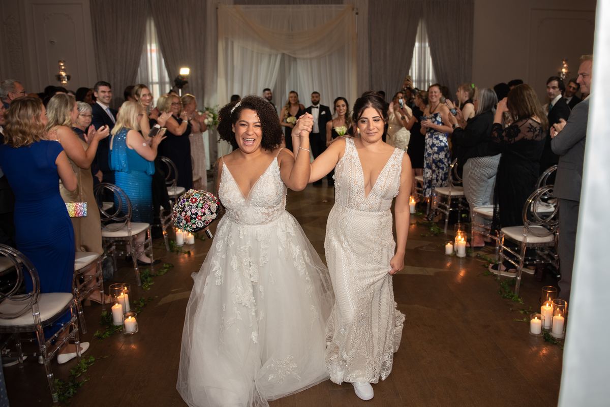 Two brides walk hand-in-hand down a candlelit aisle surrounded by cheering guests in an elegant hall.