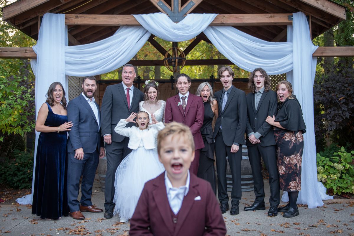 Family and friends pose joyfully under a decorated wooden gazebo during an autumn wedding celebration.