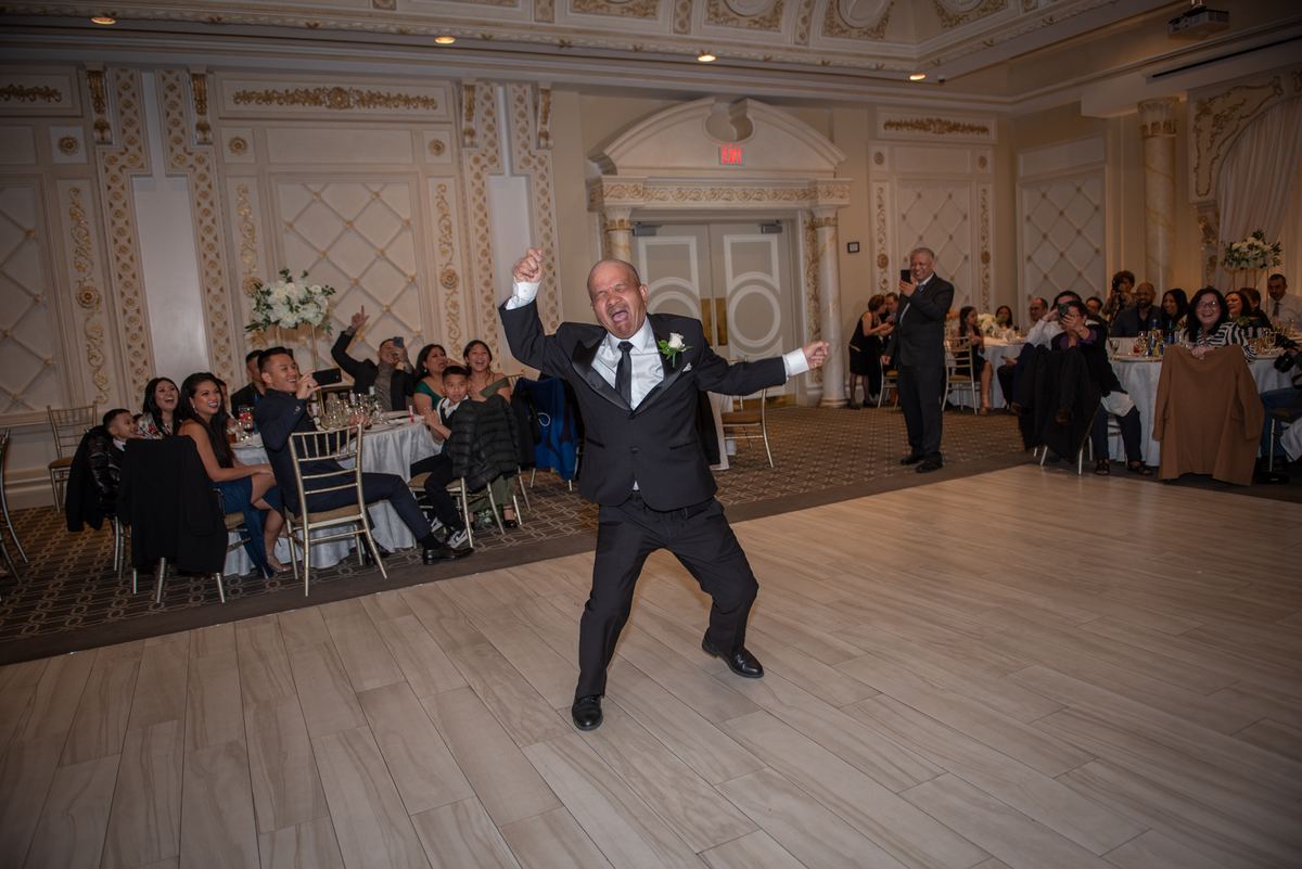 A groom in a black tuxedo dances joyfully across the ballroom floor, arms raised, as guests watch and cheer.