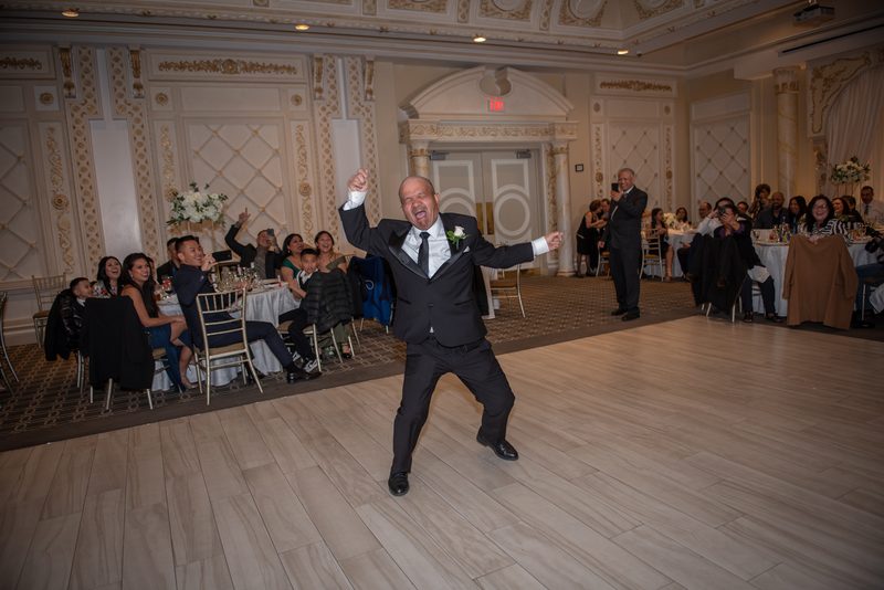 A groom in a black tuxedo dances joyfully across the ballroom floor, arms raised, as guests watch and cheer.