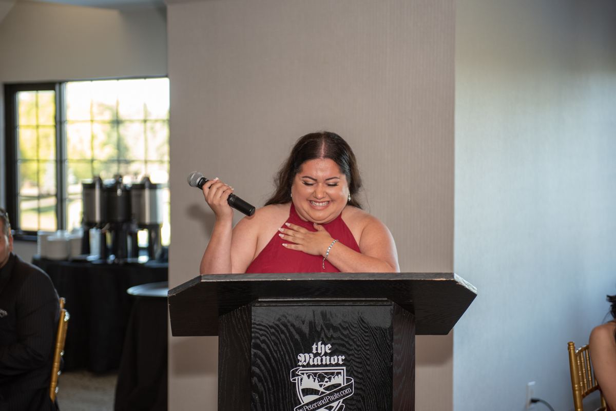 A woman in a red halter top speaks emotionally into a microphone at an indoor event with coffee stations and guests.