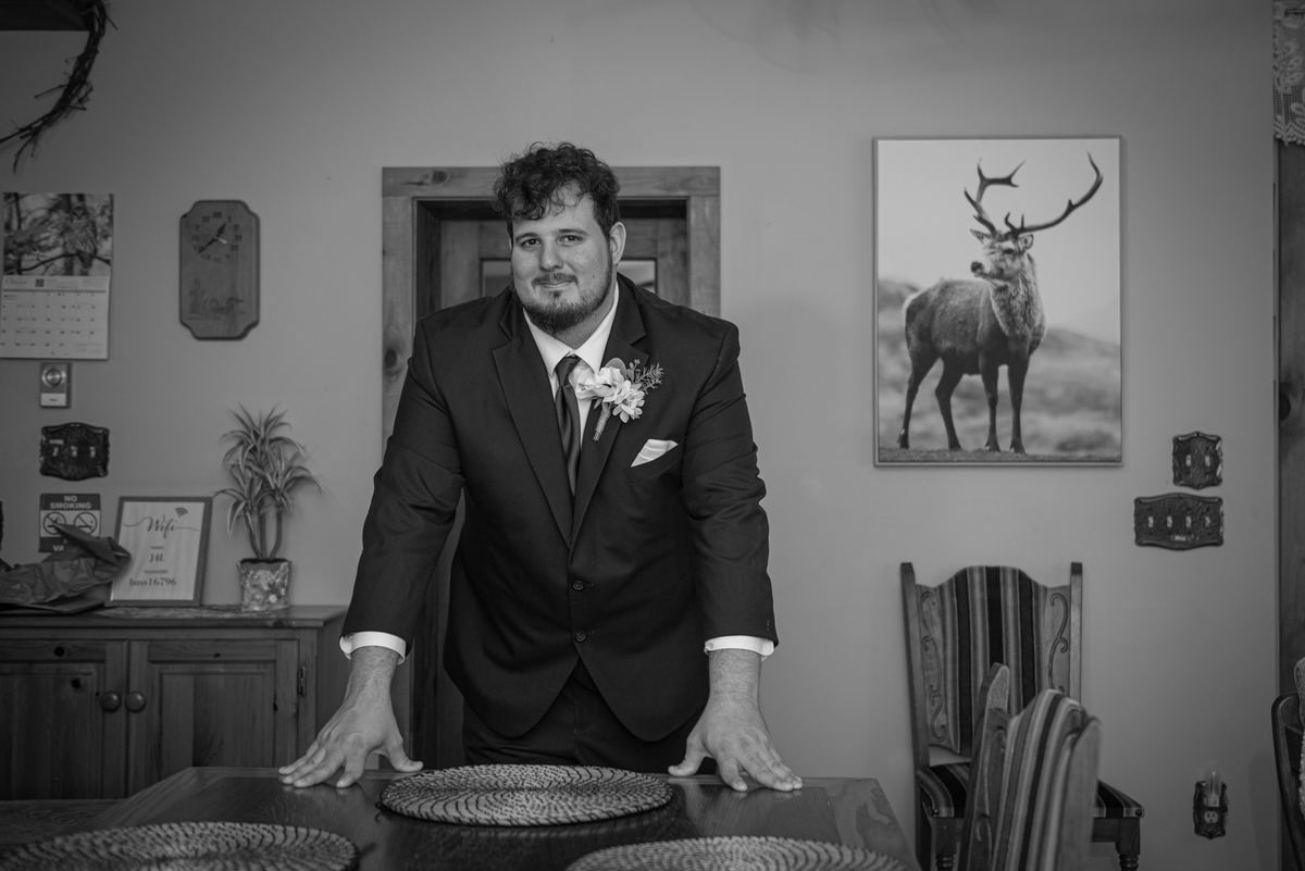 A groom in a dark suit stands at a dining table in a rustic room with wildlife art and a clock, hands resting on the surface.