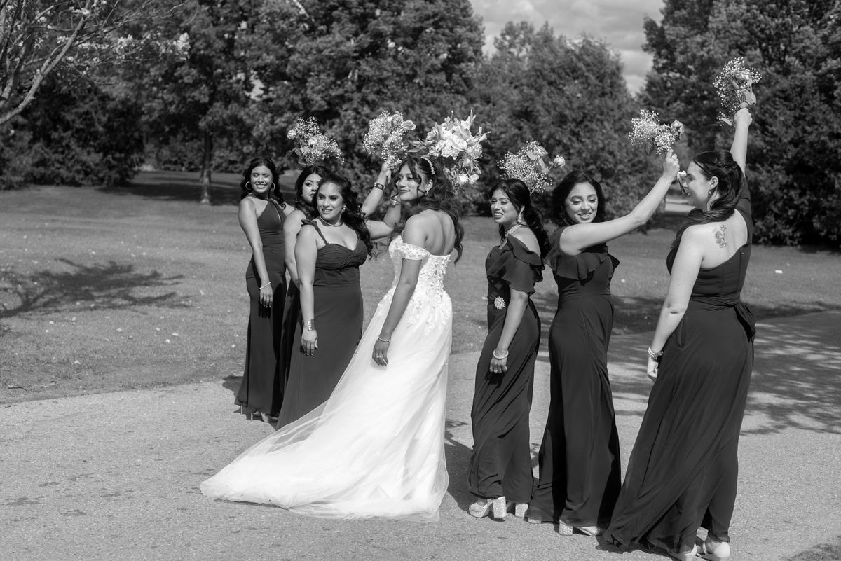 A bride and her bridesmaids stand joyfully outdoors, raising bouquets in bright sunlight filtered through trees.
