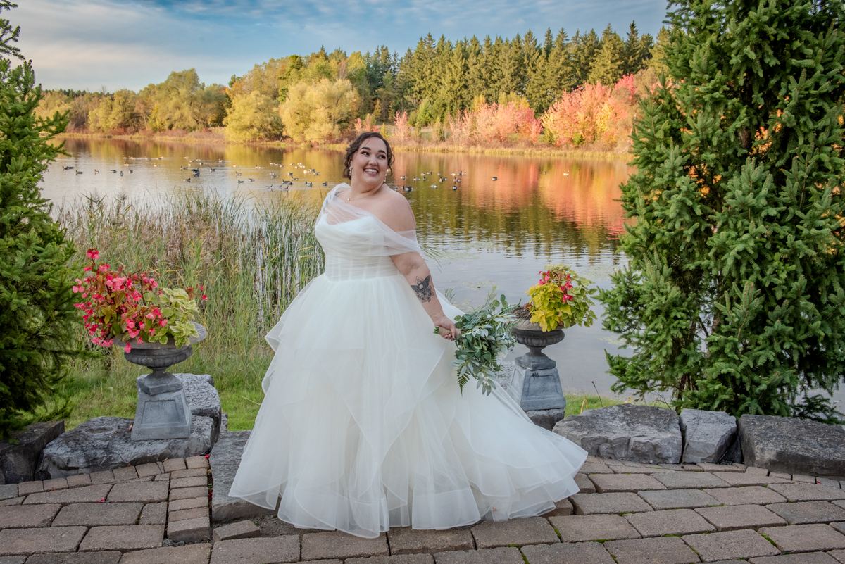 A bride stands on a stone patio by a lake, surrounded by autumn foliage and evergreen trees, holding greenery.