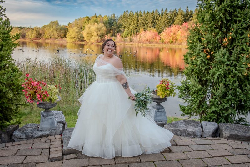 A bride stands on a stone patio by a lake, surrounded by autumn foliage and evergreen trees, holding greenery.