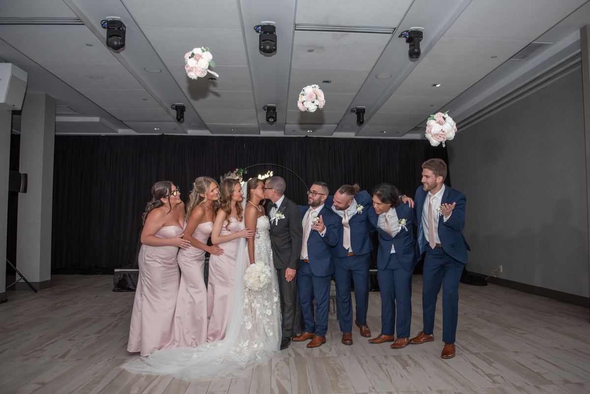 Bride and groom kiss as their smiling bridal party leans in, surrounded by soft lighting and floral decorations.