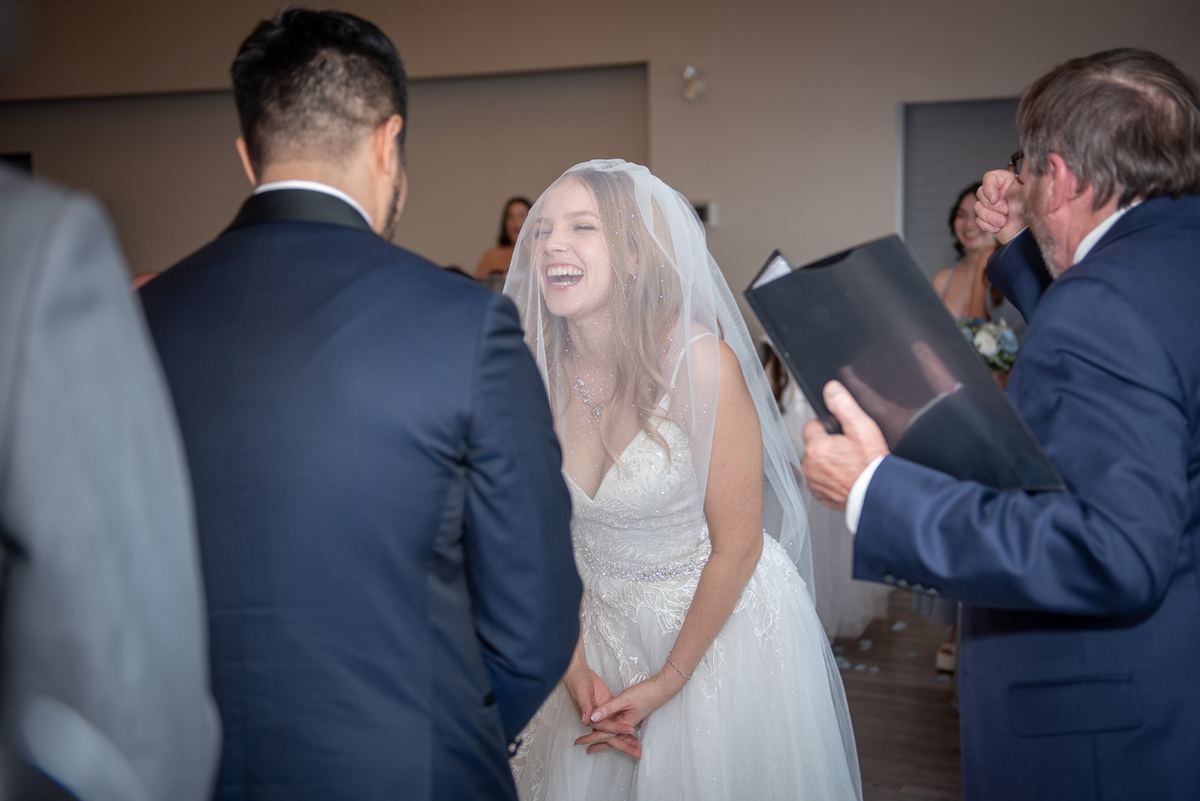The bride laughs as she stands before her groom with a man holding an open photo album, guests in the background.