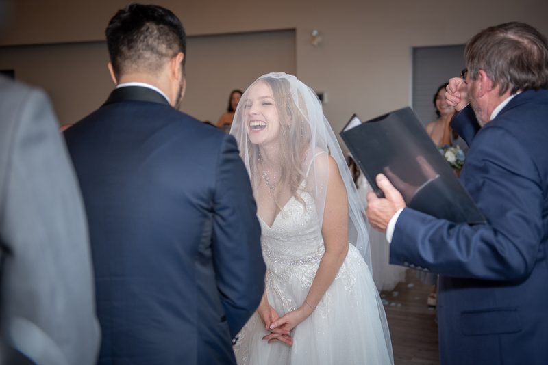 The bride laughs as she stands before her groom with a man holding an open photo album, guests in the background.