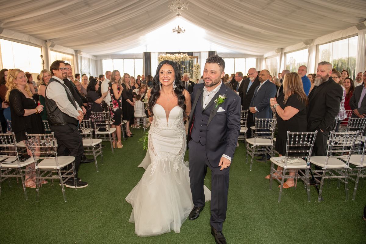 A bride and groom smile as they walk down the aisle under a draped tent, guests watching in acrylic chairs.