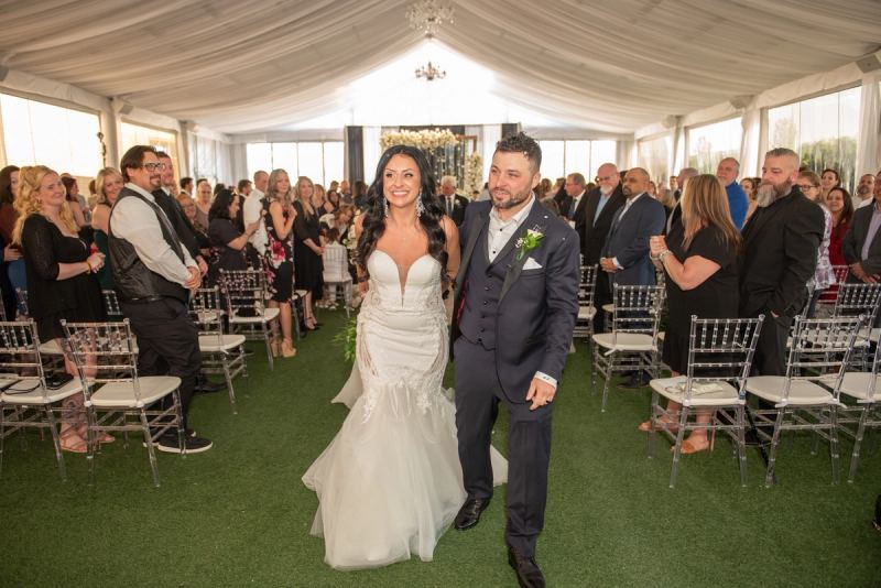 A bride and groom smile as they walk down the aisle under a draped tent, guests watching in acrylic chairs.
