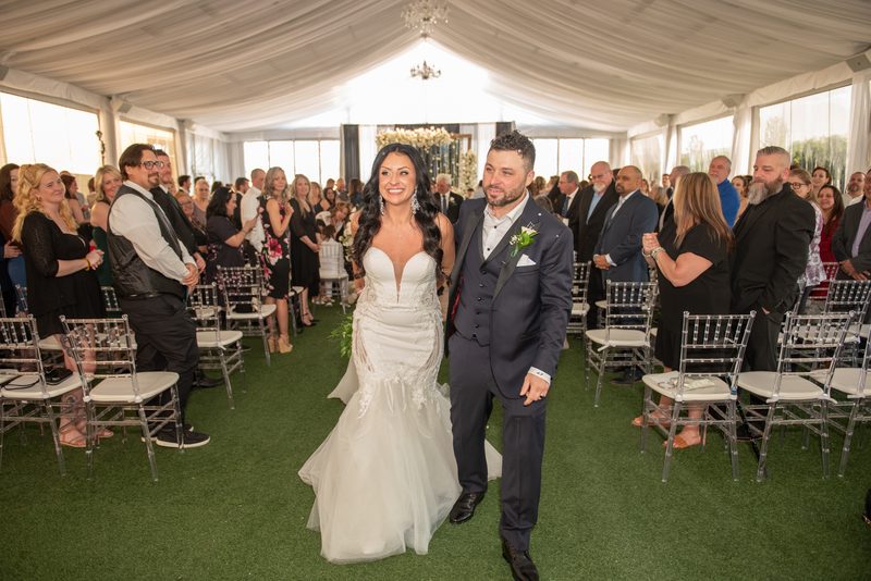 A bride and groom smile as they walk down the aisle under a draped tent, guests watching in acrylic chairs.