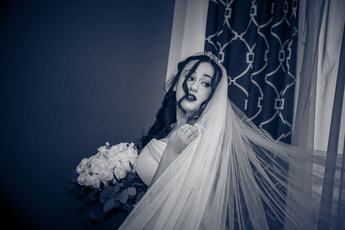 A bride in monochrome leans forward dramatically, her veil half-covered, holding white roses against a dark curtain backdrop.