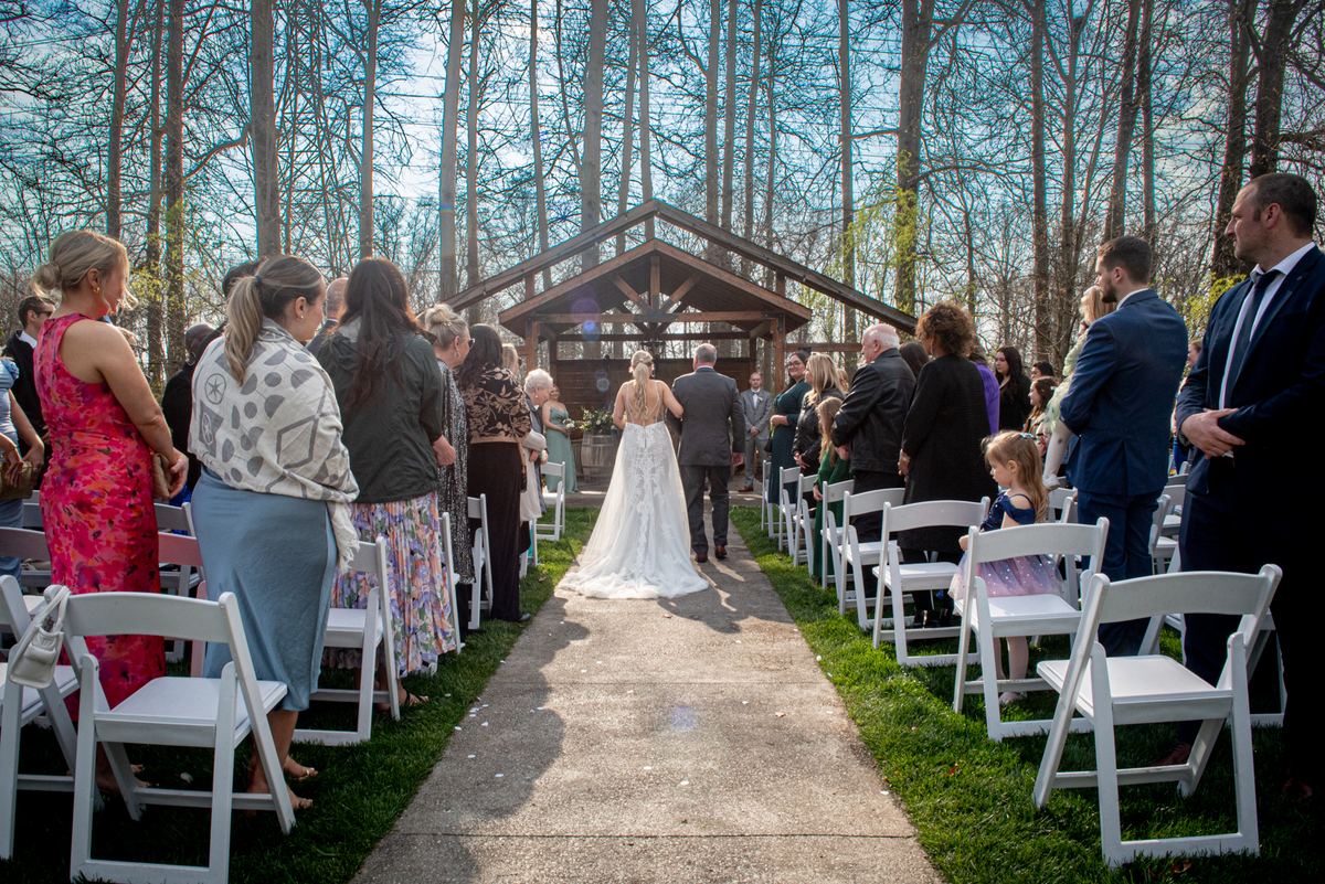 A bride in a white gown walks down an aisle toward a wooden arbor, surrounded by guests and a young girl in a sparkly dress.