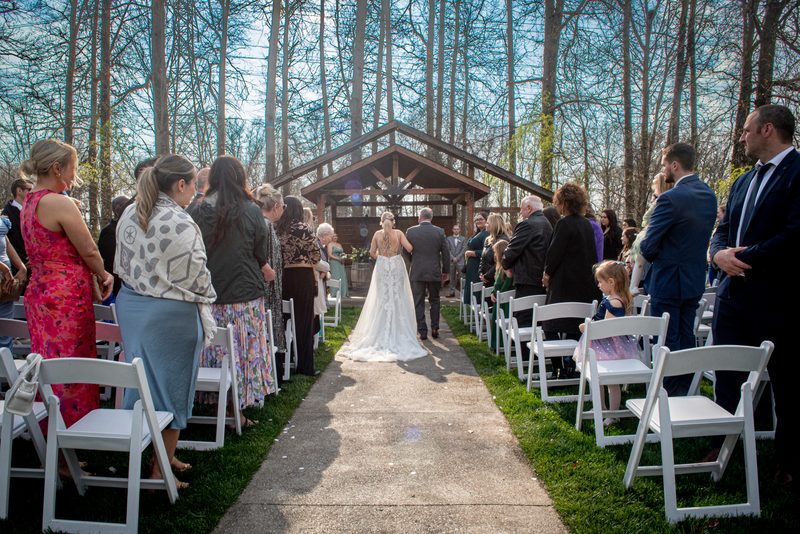 A bride in a white gown walks down an aisle toward a wooden arbor, surrounded by guests and a young girl in a sparkly dress.