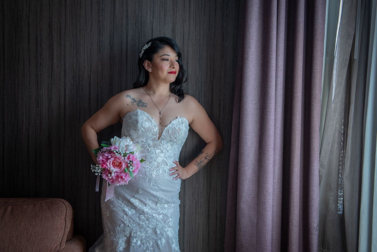 A bride in a white lace gown holds flowers beside a window, illuminated by soft light filtering through curtains.