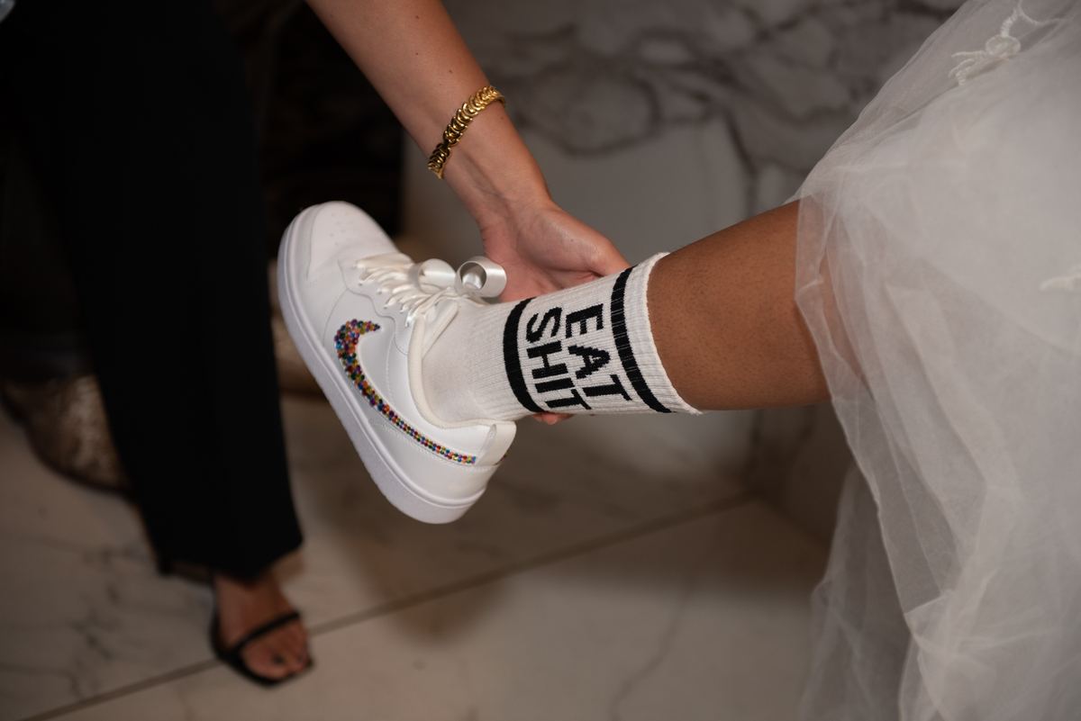 A bride in a white gown has her foot lifted as someone helps put on a bejeweled sneaker, contrasting with bold socks.