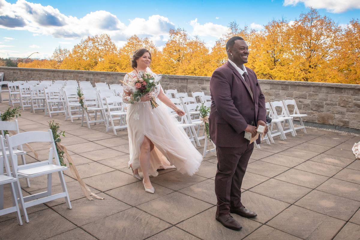 A bride in a white gown walks down the aisle toward her groom as autumn trees and a blue sky frame the stone patio.