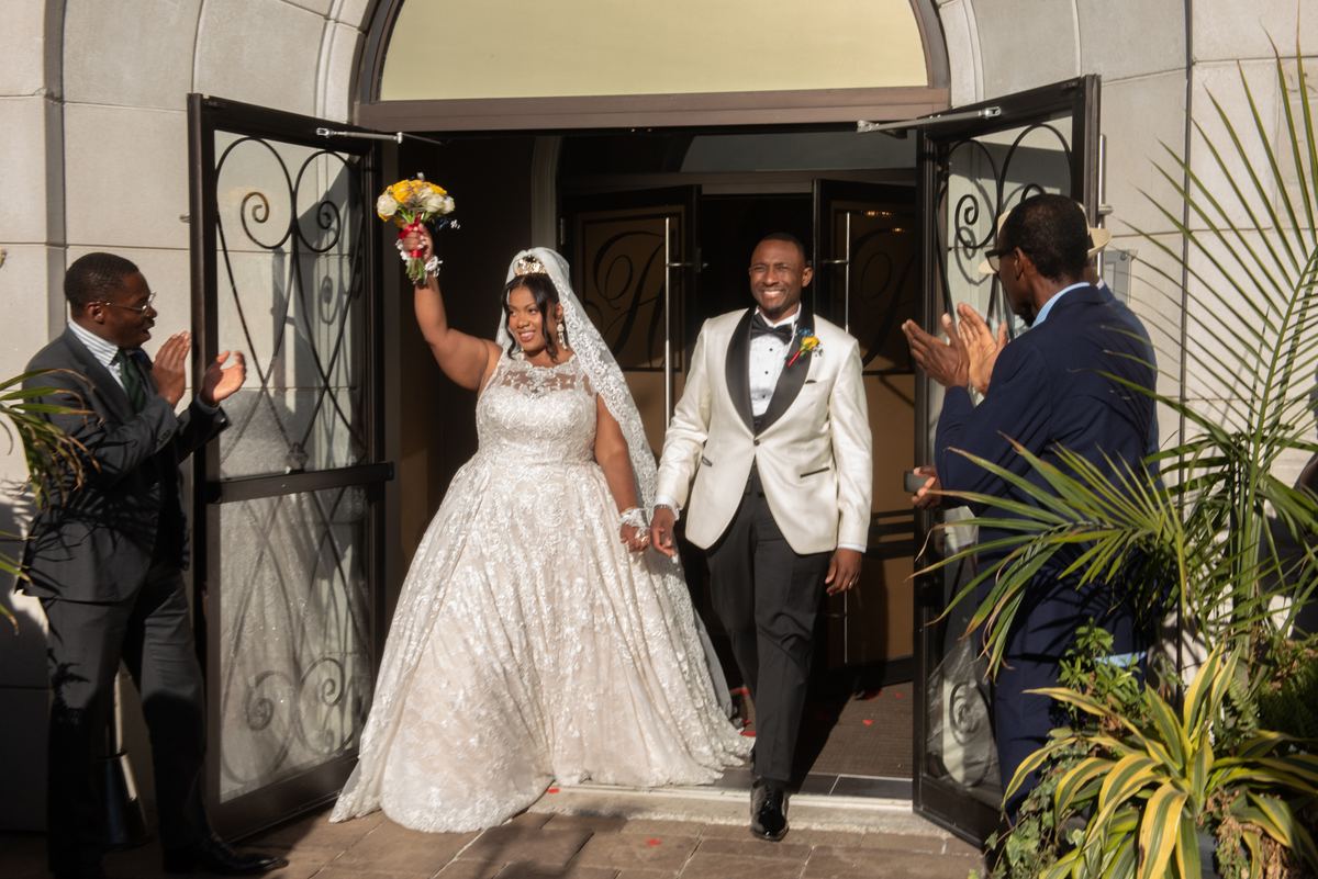 A smiling bride and groom exit through ornate doors, waving and smiling back at applauding guests in bright sunlight.