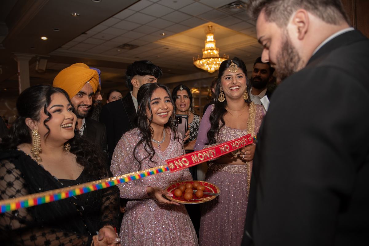 A bride in pink holds sweets as a guest approaches a ceremonial ribbon, celebrating indoors with festive guests.