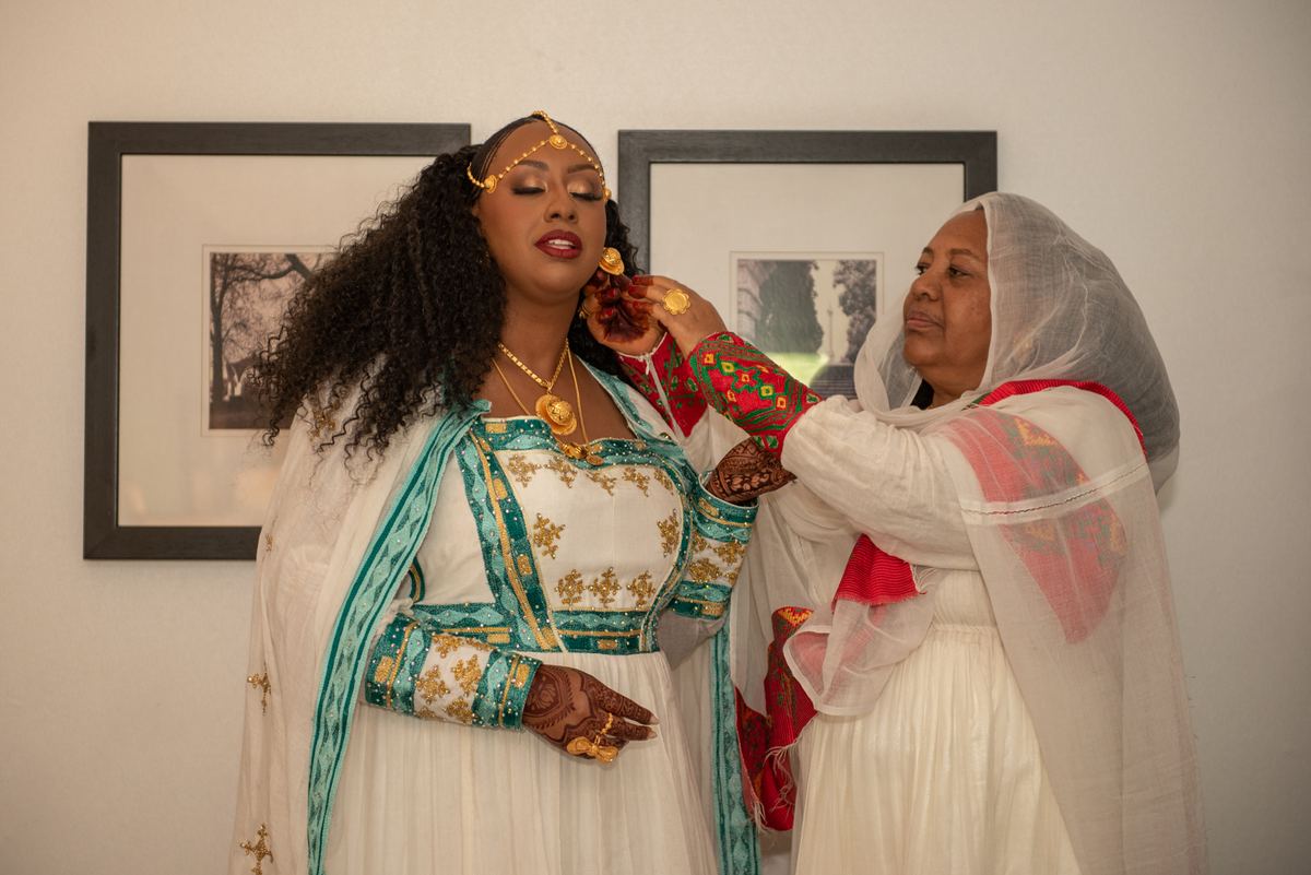 A young woman in a white and teal gown receives an ornament from an older woman during a cultural ceremony.