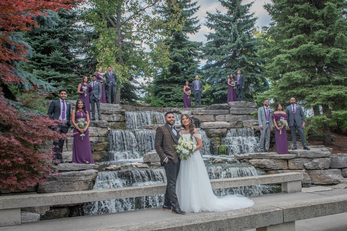 A wedding party poses in front of a stone waterfall, with the bride and groom centered, dressed in purple gowns.