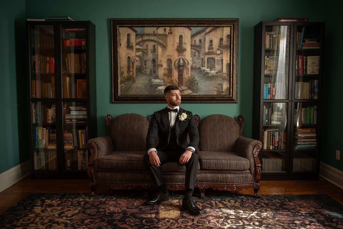 A groom in a tuxedo sits thoughtfully on an ornate sofa in a richly decorated room with bookshelves.