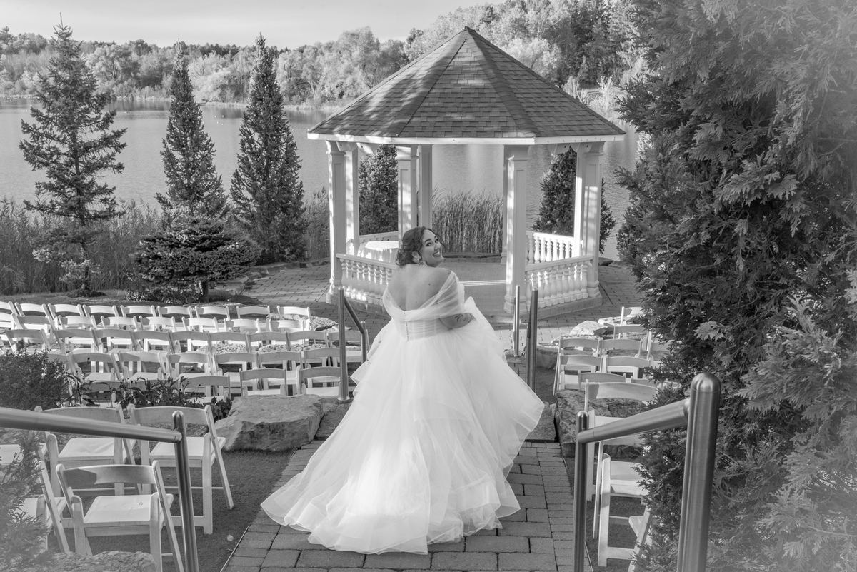 A bride in a white gown smiles on a stone path near empty chairs and a gazebo by a serene lake, framed by evergreens.