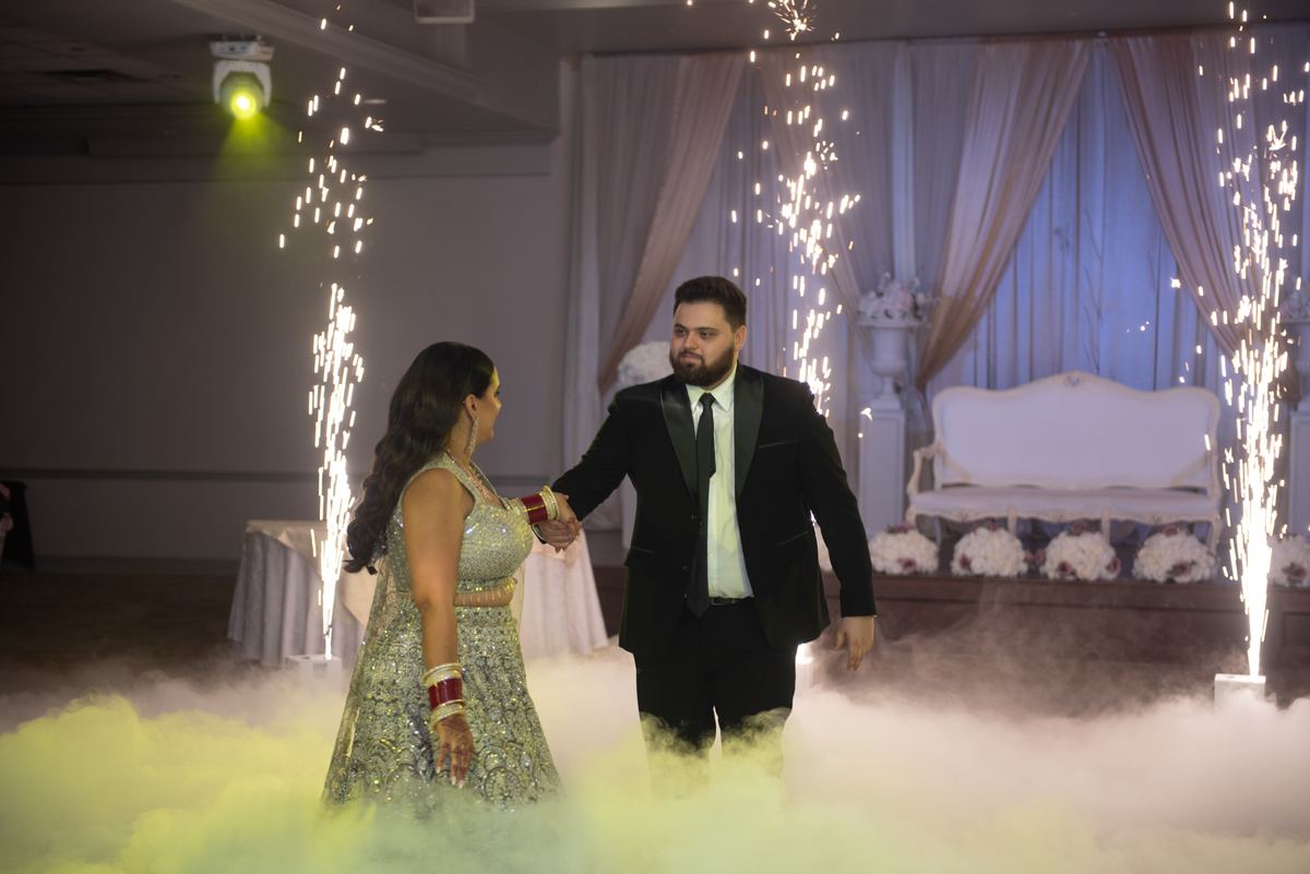 A couple in formal attire stands center stage, illuminated by green light and surrounded by fog and sparklers.