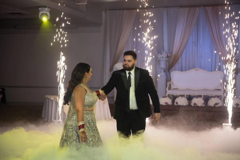 A couple in formal attire stands center stage, illuminated by green light and surrounded by fog and sparklers.