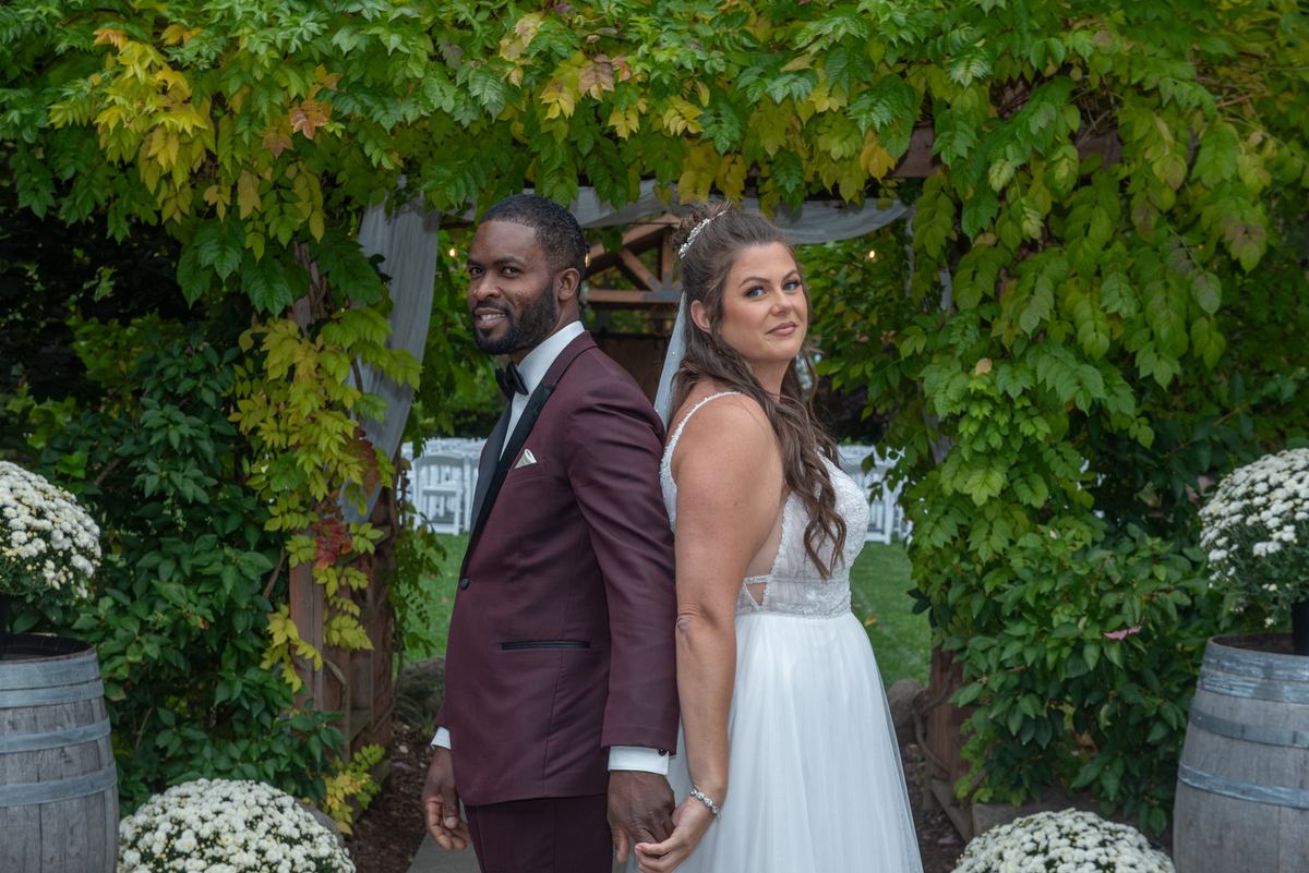 Alexandria and Derado stand back-to-back under a lush green arbor, smiling at the camera in their wedding attire.