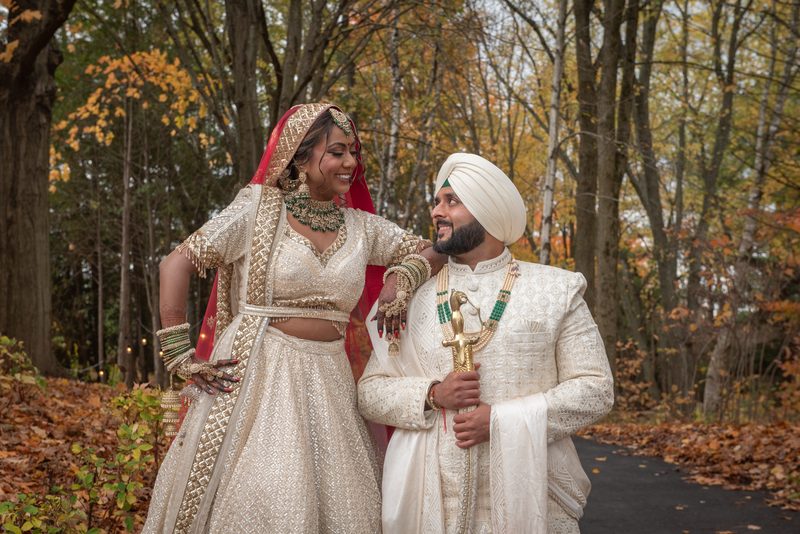 A smiling bride and groom in traditional Indian attire walk through an autumn forest, light filtering through trees.