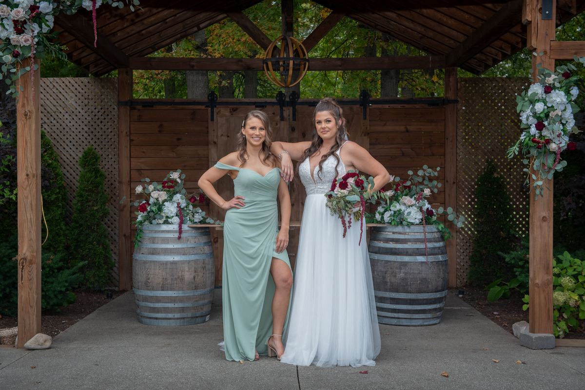Two brides stand under a wooden arbor decorated with flowers, smiling beside wine barrels adorned with floral arrangements.