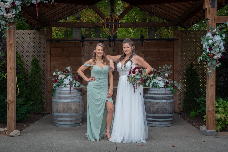 Two brides stand under a wooden arbor decorated with flowers, smiling beside wine barrels adorned with floral arrangements.