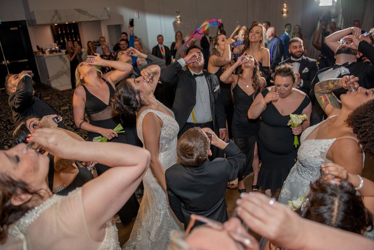 Guests in formal attire raise glasses and toast at a wedding reception, illuminated by warm lights and festive decorations.