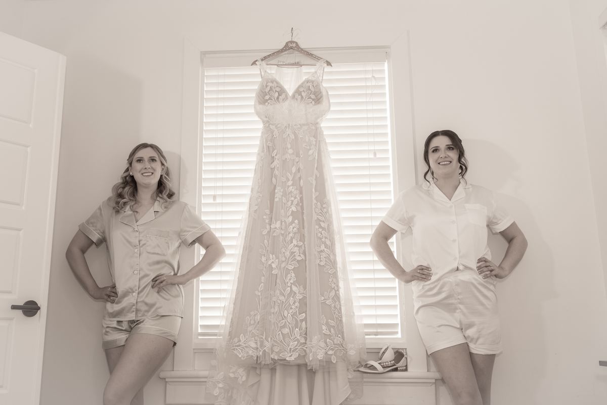 Two bridesmaids stand beside a window, smiling as they flank Amanda's stunning wedding gown hanging on a hanger.