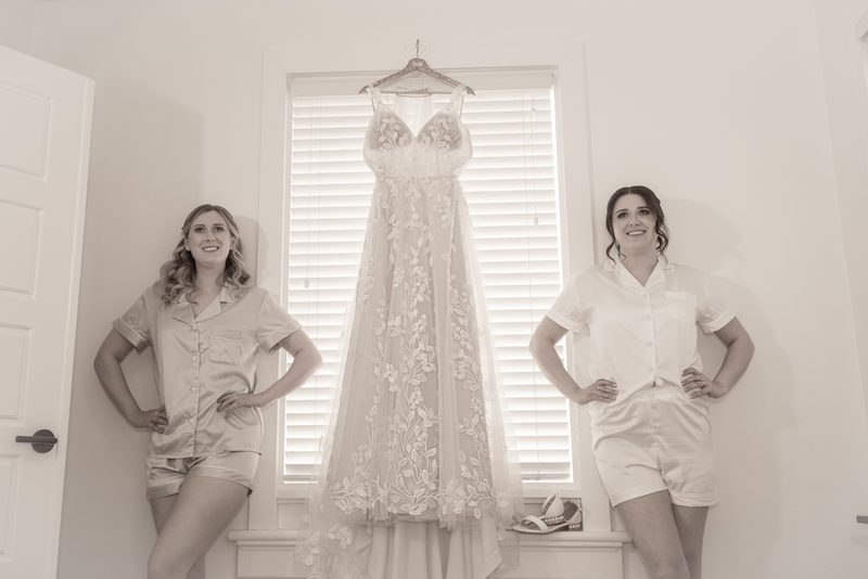 Two bridesmaids stand beside a window, smiling as they flank Amanda's stunning wedding gown hanging on a hanger.