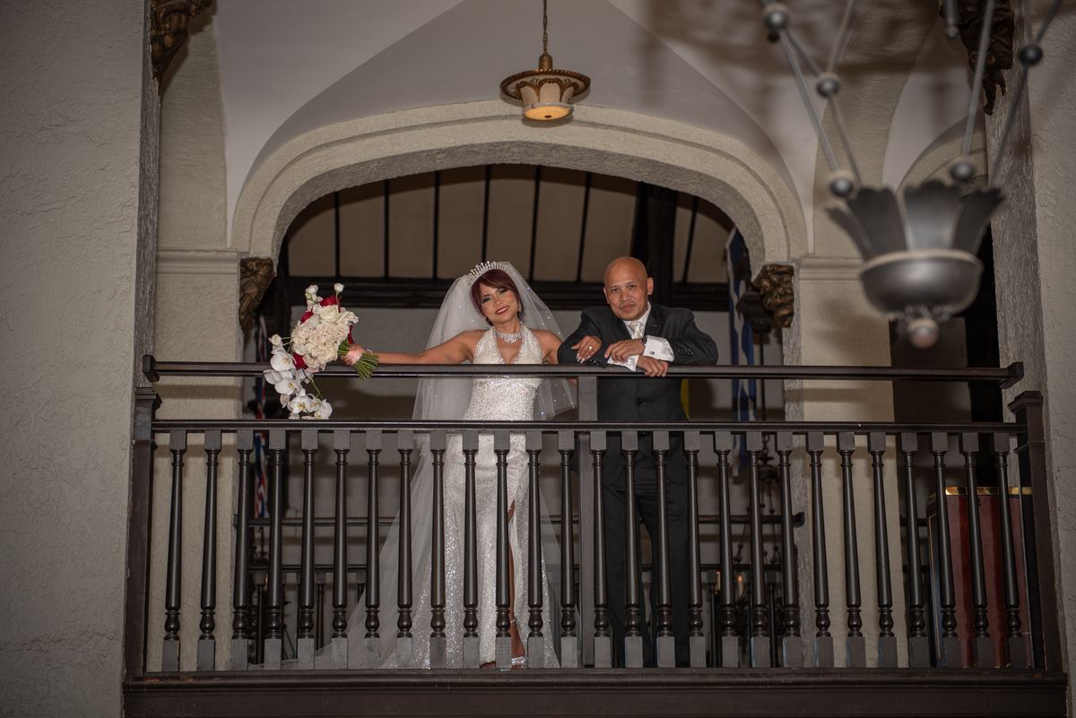 A bride and groom smile on an ornate balcony, illuminated by hanging lights against arched architecture.