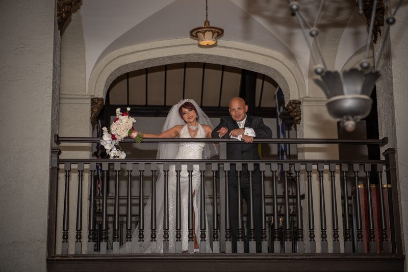 A bride and groom smile on an ornate balcony, illuminated by hanging lights against arched architecture.