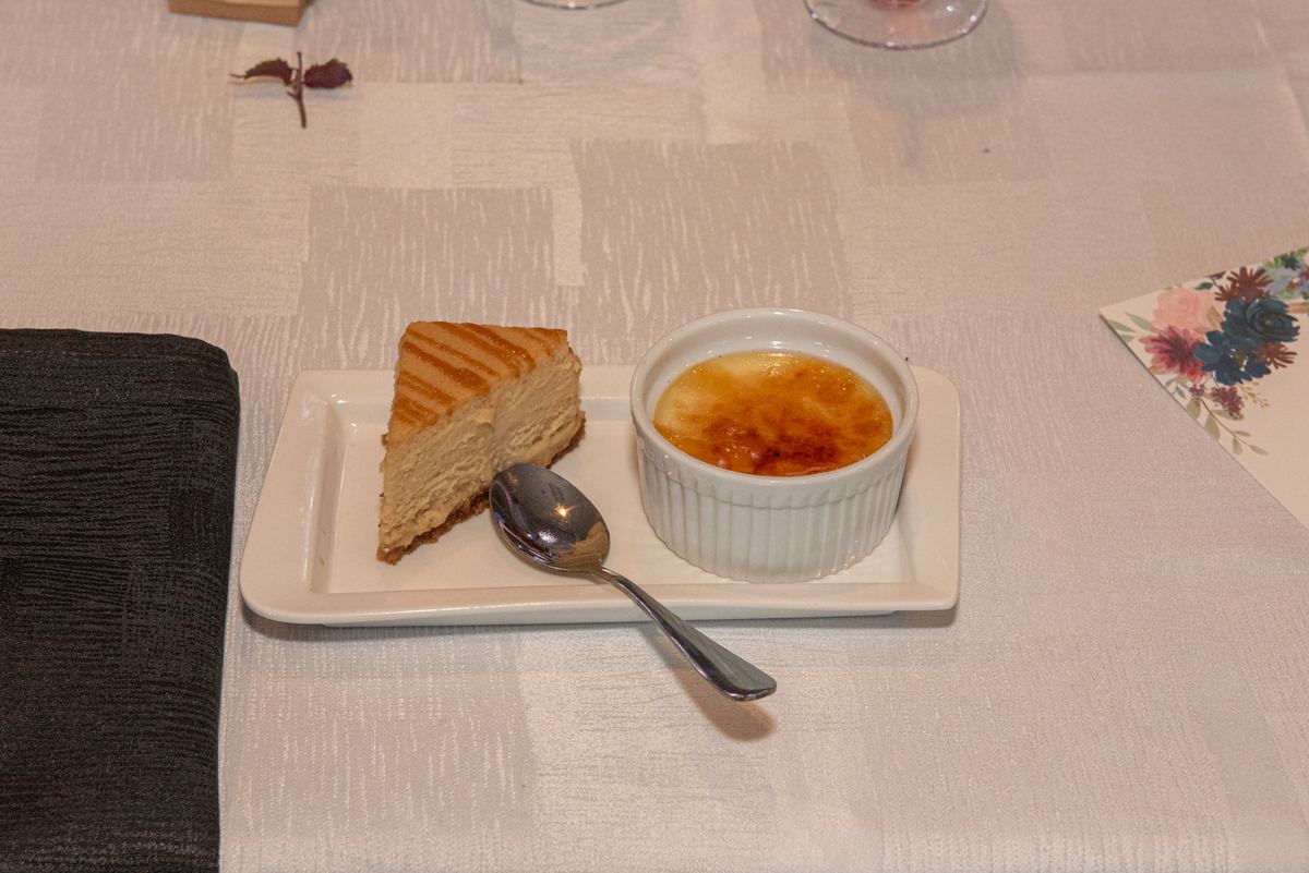 A white plate holds a slice of grilled cheesecake and crème brûlée with a spoon beside it, suggesting a quiet dessert moment.