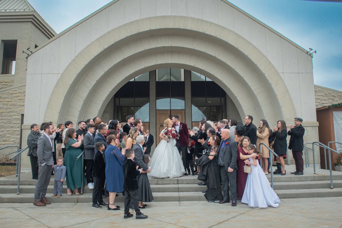 A bride and groom kiss on church steps surrounded by cheering guests in formal wear under an overcast sky.