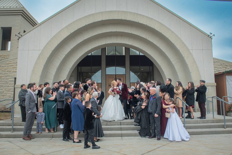 A bride and groom kiss on church steps surrounded by cheering guests in formal wear under an overcast sky.