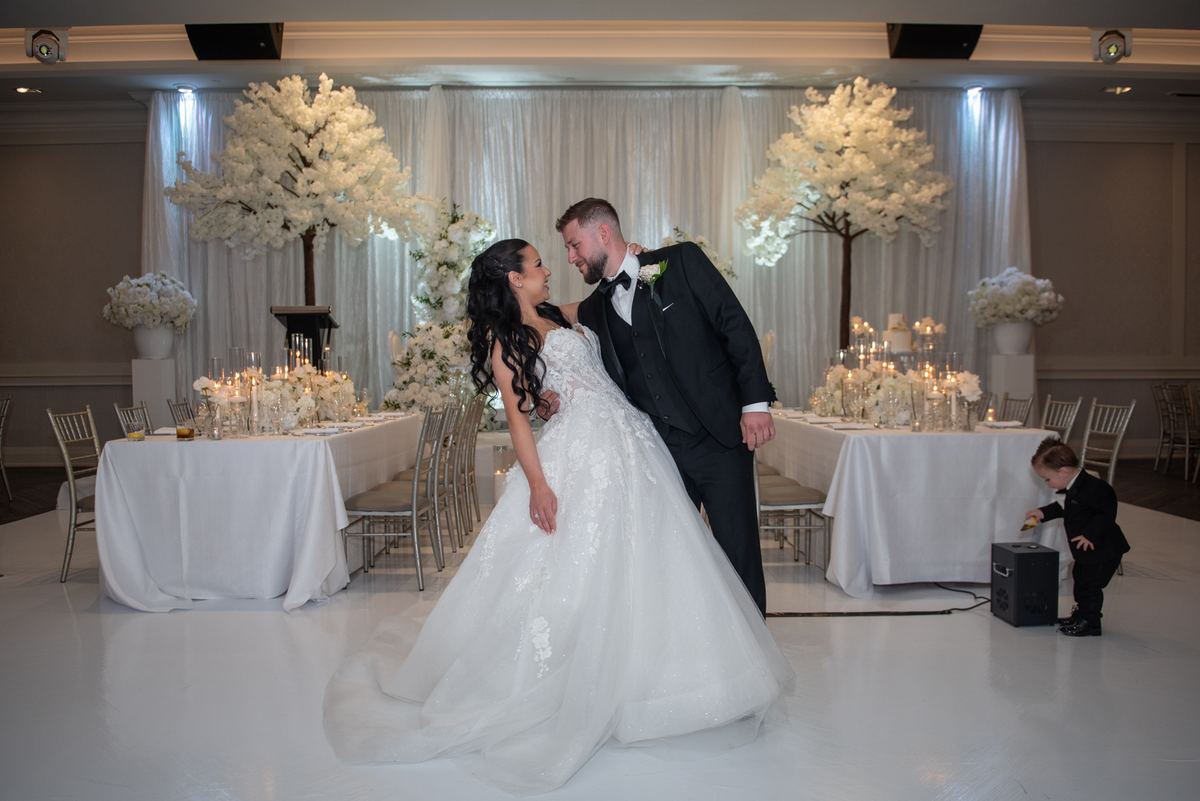 Bride and groom kiss on the dance floor as soft lighting illuminates their white-themed celebration.