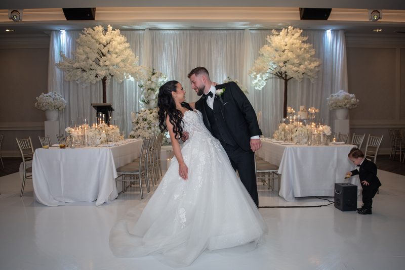 Bride and groom kiss on the dance floor as soft lighting illuminates their white-themed celebration.