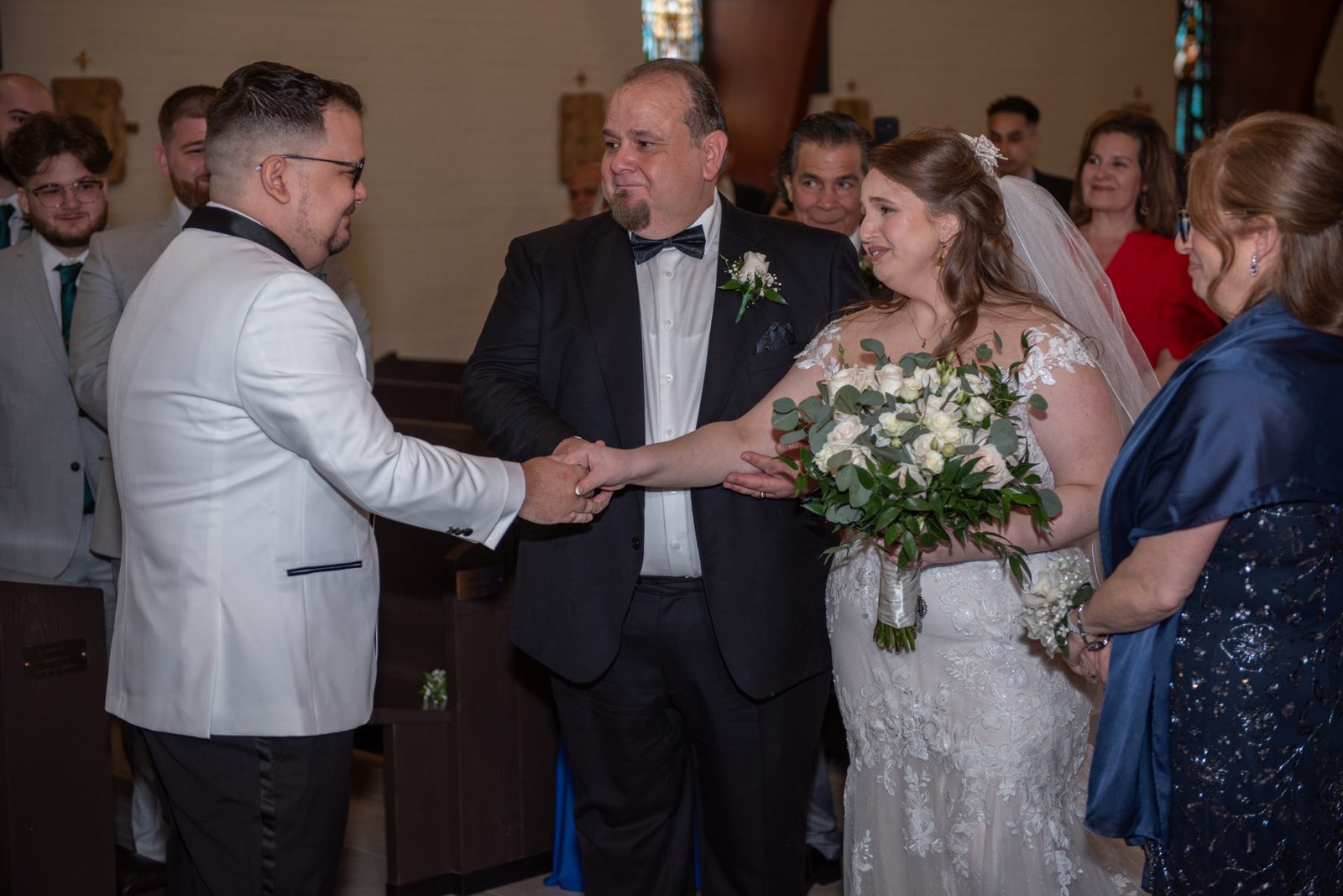 A bride and groom exchange hands with the officiant during a wedding ceremony in a church, surrounded by guests.