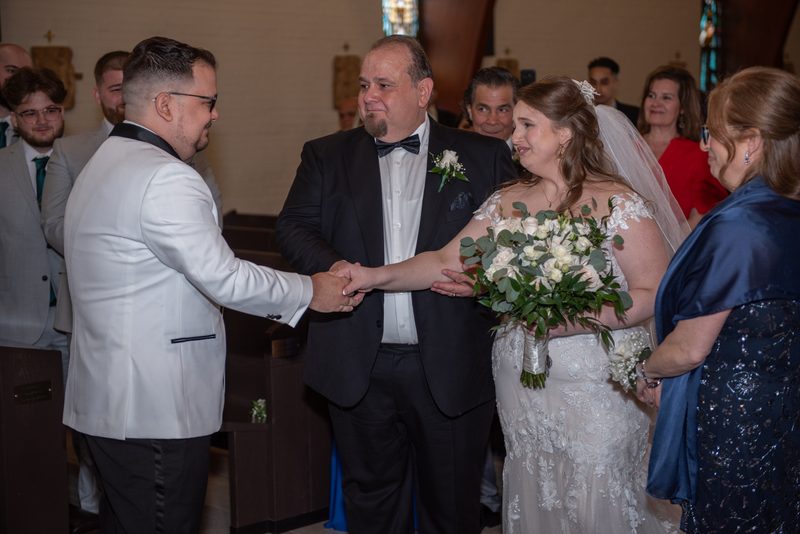 A bride and groom exchange hands with the officiant during a wedding ceremony in a church, surrounded by guests.