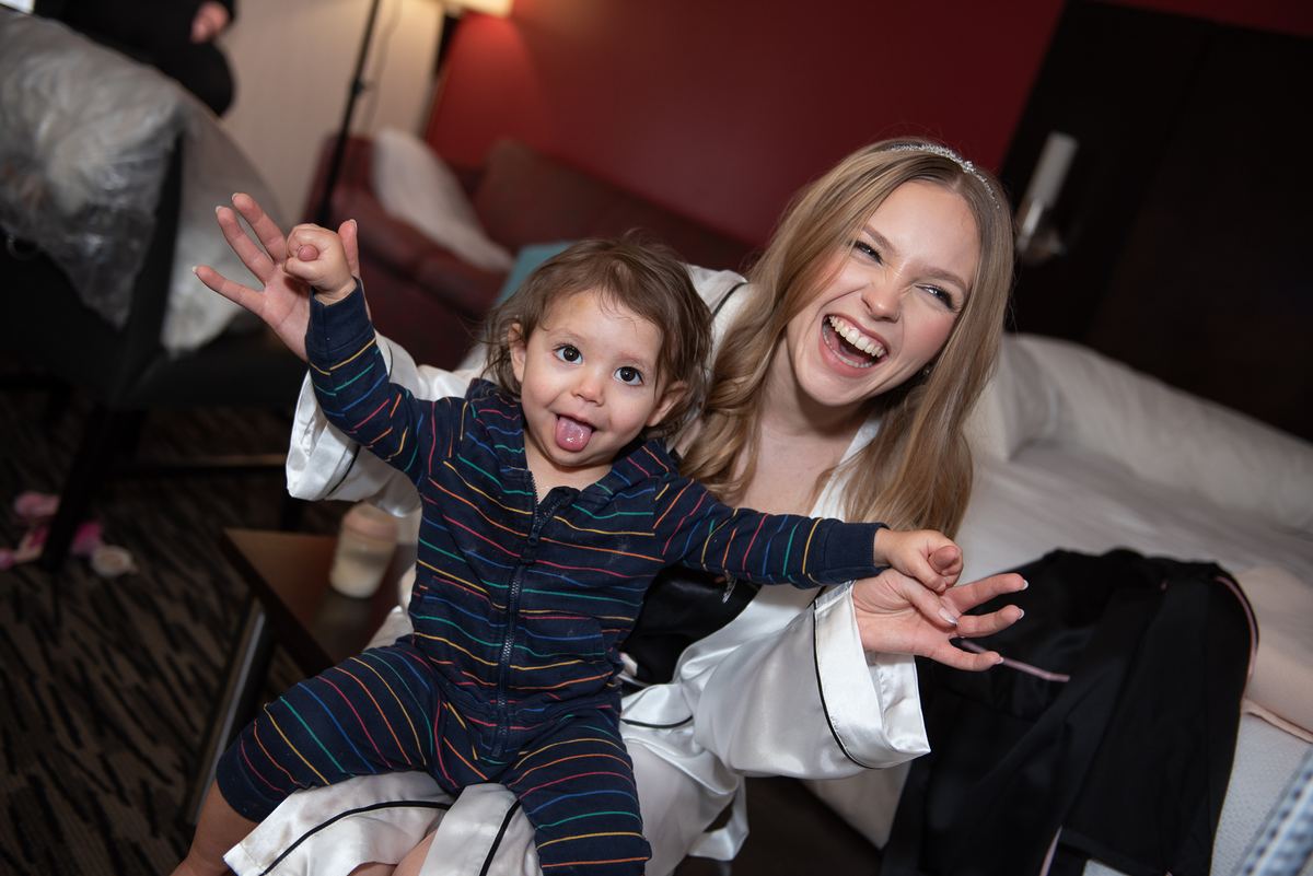 A woman in a satin robe laughs with a toddler who sticks out his tongue and gestures playfully, creating an intimate moment.