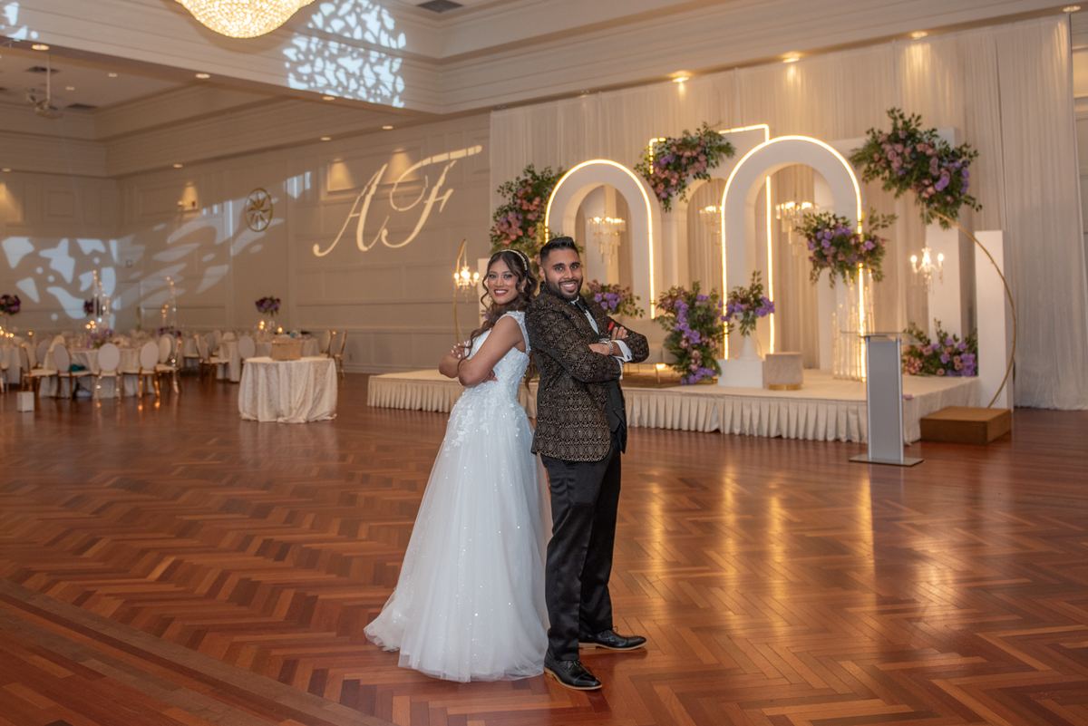 A smiling bride and groom pose on the dance floor of an elegantly decorated wedding hall with warm lighting.