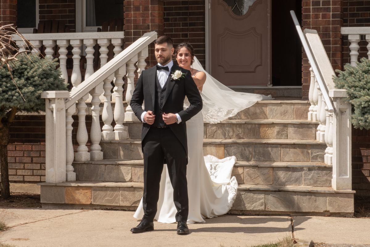 A bride and groom stand on stone steps, the sunlight highlighting textures of the brick house and white balustrade.