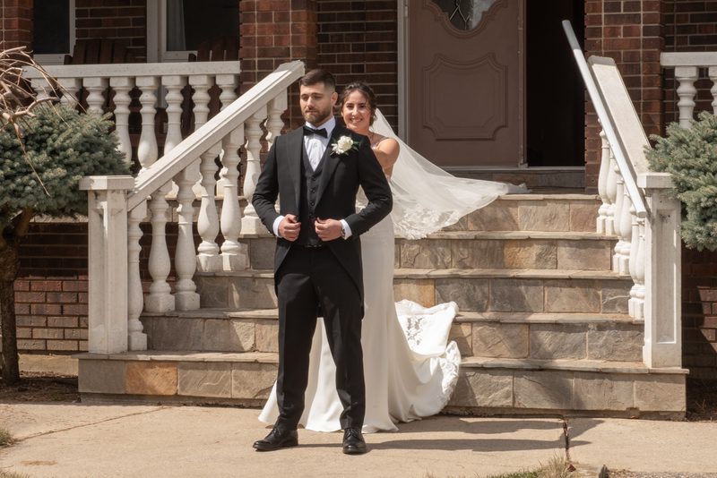 A bride and groom stand on stone steps, the sunlight highlighting textures of the brick house and white balustrade.