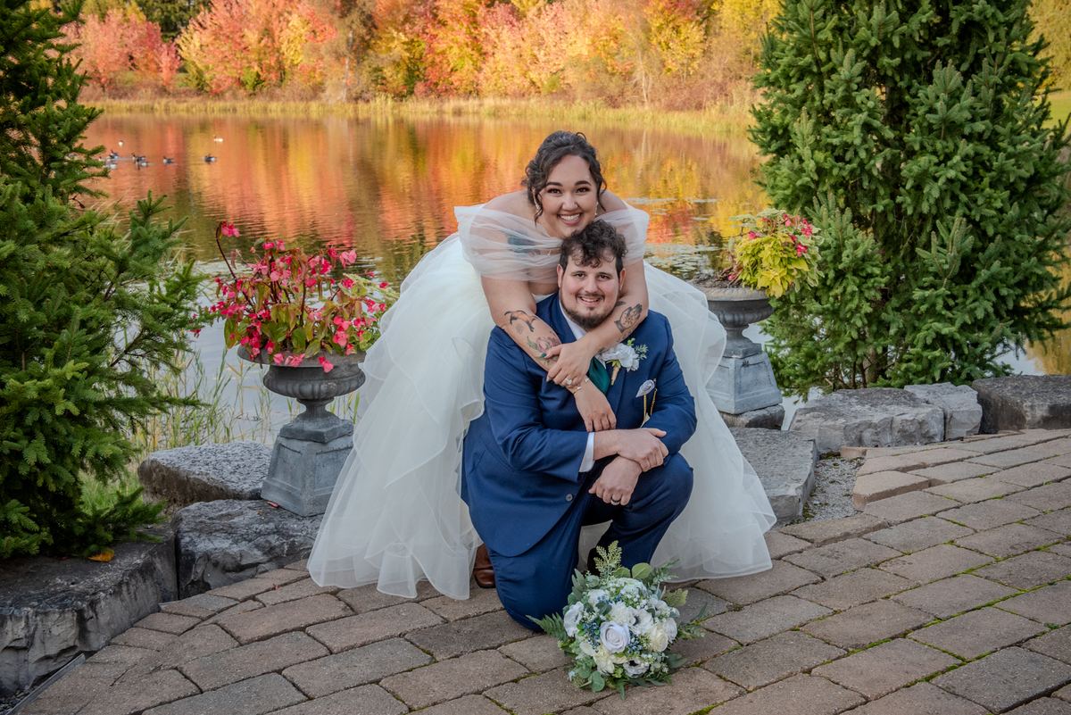 A bride perched on her groom's shoulders by a pond, both dressed in wedding attire, bathed in autumn's golden light.